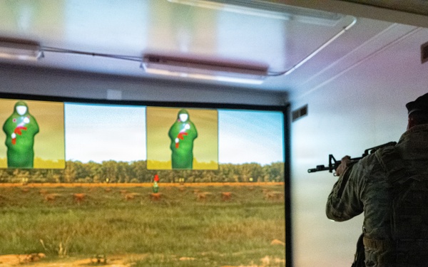 A soldier in full combat gear and camouflage uniform is using a firearms simulation training system, aiming a rifle at projected targets on a large screen. The simulation displays a realistic outdoor range environment with green humanoid silhouette targets showing red hit markers. Multiple training rifles are laid out on the floor behind the soldier, suggesting a group training session.
