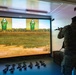 A soldier in full combat gear and camouflage uniform is using a firearms simulation training system, aiming a rifle at projected targets on a large screen. The simulation displays a realistic outdoor range environment with green humanoid silhouette targets showing red hit markers. Multiple training rifles are laid out on the floor behind the soldier, suggesting a group training session.