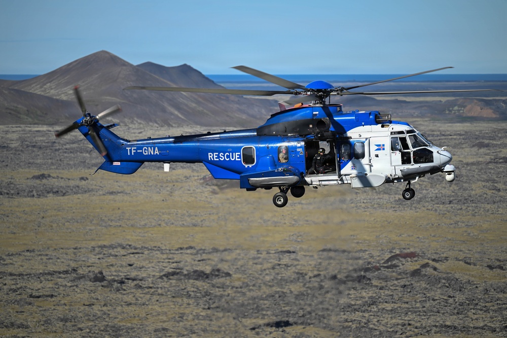 A blue and white search and rescue helicopter with registration TF-GNA is flying low over a barren, volcanic landscape. The helicopter bears the word 'RESCUE' on its fuselage and displays what appears to be an Icelandic flag emblem, suggesting it belongs to the Icelandic Coast Guard or a related rescue service. The terrain and volcanic mountains in the background are consistent with Iceland's landscape.