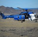 A blue and white search and rescue helicopter with registration TF-GNA is flying low over a barren, volcanic landscape. The helicopter bears the word 'RESCUE' on its fuselage and displays what appears to be an Icelandic flag emblem, suggesting it belongs to the Icelandic Coast Guard or a related rescue service. The terrain and volcanic mountains in the background are consistent with Iceland's landscape.