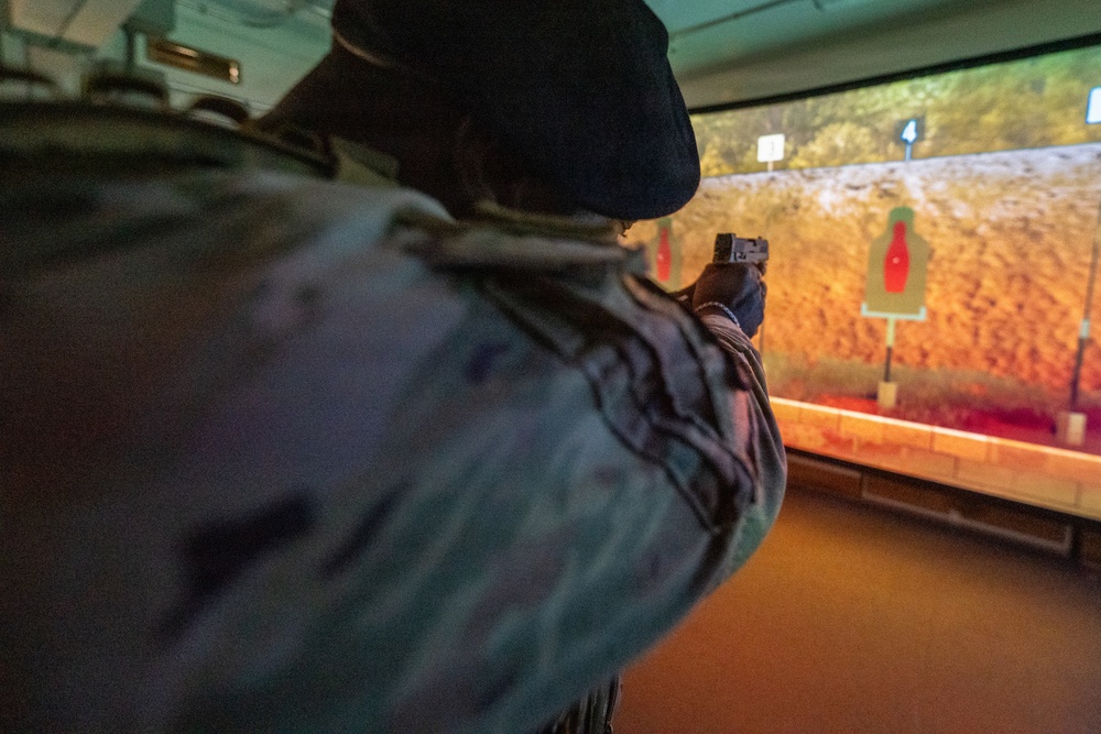 A person in military camouflage uniform and black beret is aiming a pistol at a shooting range simulator. The projected screen displays a rocky outdoor environment with red bottle-shaped targets. This appears to be an indoor virtual marksmanship training facility.