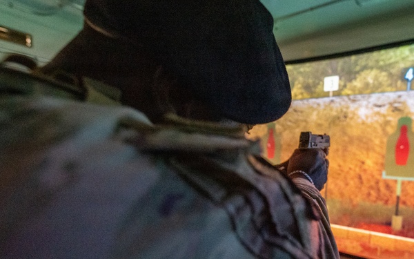 A person in military camouflage uniform and black beret is aiming a pistol at a shooting range simulator. The projected screen displays a rocky outdoor environment with red bottle-shaped targets. This appears to be an indoor virtual marksmanship training facility.
