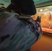 A person in military camouflage uniform and black beret is aiming a pistol at a shooting range simulator. The projected screen displays a rocky outdoor environment with red bottle-shaped targets. This appears to be an indoor virtual marksmanship training facility.