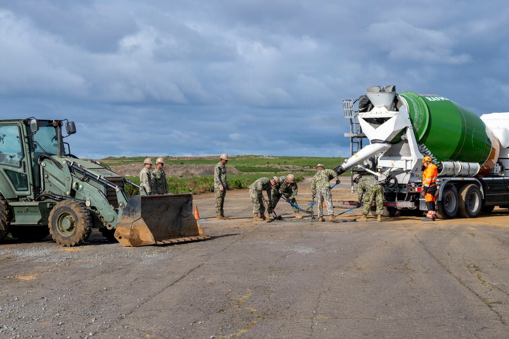 Military personnel in Navy working uniforms are conducting a concrete pouring operation on what appears to be a runway or tarmac surface. A green and white concrete mixer truck is dispensing concrete while several sailors work to spread and manage the pour using tools. A military front-end loader is positioned nearby on the left side of the scene.