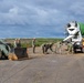 Military personnel in Navy working uniforms are conducting a concrete pouring operation on what appears to be a runway or tarmac surface. A green and white concrete mixer truck is dispensing concrete while several sailors work to spread and manage the pour using tools. A military front-end loader is positioned nearby on the left side of the scene.