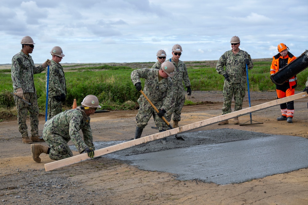 A group of military personnel in U.S. Navy digital camouflage uniforms and tan hard hats are performing concrete repair work on a damaged road or surface. They are using tools including rakes, shovels, and a wooden screed board to level fresh concrete over a patched area. One civilian worker in high-visibility orange gear is also present, assisting with the operation.