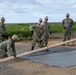 A group of military personnel in U.S. Navy digital camouflage uniforms and tan hard hats are performing concrete repair work on a damaged road or surface. They are using tools including rakes, shovels, and a wooden screed board to level fresh concrete over a patched area. One civilian worker in high-visibility orange gear is also present, assisting with the operation.