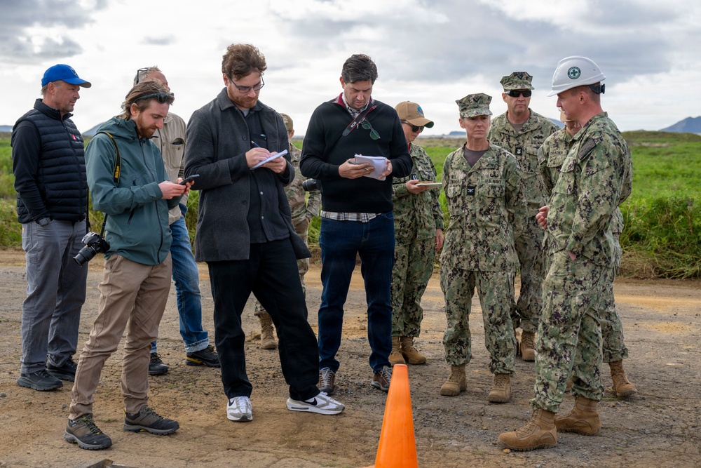 A group of civilians and U.S. Navy personnel are gathered outdoors on a dirt surface, appearing to be engaged in a briefing or site inspection. Several civilians are taking notes on notepads while military personnel in NWU Type III camouflage uniforms, including one wearing a white hard hat, appear to be conducting or participating in the meeting. An orange traffic cone is visible in the foreground, suggesting a marked or controlled area.