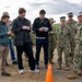 A group of civilians and U.S. Navy personnel are gathered outdoors on a dirt surface, appearing to be engaged in a briefing or site inspection. Several civilians are taking notes on notepads while military personnel in NWU Type III camouflage uniforms, including one wearing a white hard hat, appear to be conducting or participating in the meeting. An orange traffic cone is visible in the foreground, suggesting a marked or controlled area.
