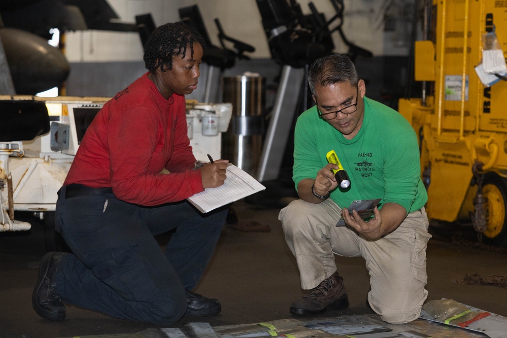 Two individuals are crouched on a floor in what appears to be a hangar or industrial workspace, conducting an inspection or inventory. The person on the left in a red shirt is taking notes on a clipboard, while the person on the right in a green shirt is using a yellow flashlight to examine items on the floor. Various equipment and machinery are visible in the background.
