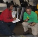 Two individuals are crouched on a floor in what appears to be a hangar or industrial workspace, conducting an inspection or inventory. The person on the left in a red shirt is taking notes on a clipboard, while the person on the right in a green shirt is using a yellow flashlight to examine items on the floor. Various equipment and machinery are visible in the background.