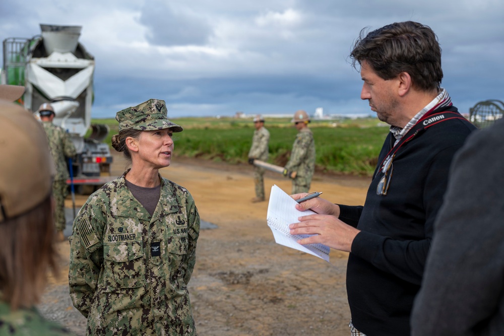 A U.S. Navy service member with the name tape 'SLOOTMAKER' is being interviewed by a civilian journalist who is holding a notepad and pen. The setting appears to be an outdoor construction or infrastructure site with military personnel and heavy equipment visible in the background. The overcast sky and muddy terrain suggest an active field operation.