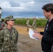 A U.S. Navy service member with the name tape 'SLOOTMAKER' is being interviewed by a civilian journalist who is holding a notepad and pen. The setting appears to be an outdoor construction or infrastructure site with military personnel and heavy equipment visible in the background. The overcast sky and muddy terrain suggest an active field operation.