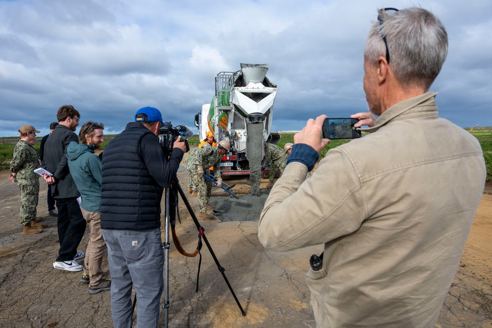 A group of observers, including media personnel and military personnel in Navy Working Uniform (NWU), are documenting a concrete pouring operation at what appears to be an airfield or runway repair site. A cement mixer truck is actively discharging concrete while workers in safety gear manage the pour. The scene suggests a military construction or infrastructure repair demonstration being covered by press.
