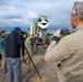 A group of observers, including media personnel and military personnel in Navy Working Uniform (NWU), are documenting a concrete pouring operation at what appears to be an airfield or runway repair site. A cement mixer truck is actively discharging concrete while workers in safety gear manage the pour. The scene suggests a military construction or infrastructure repair demonstration being covered by press.