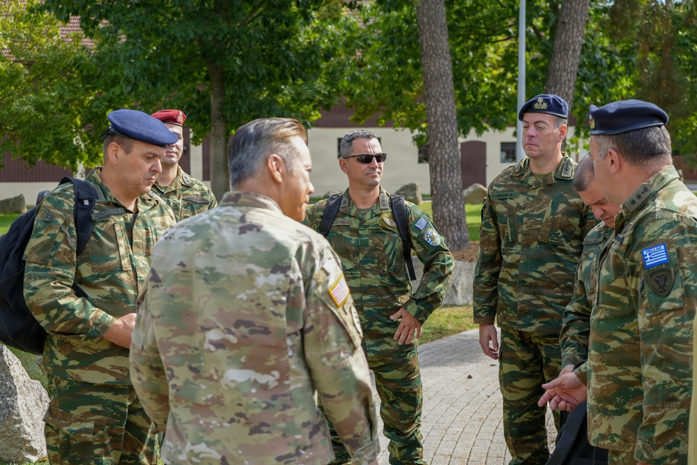 A group of military personnel from at least two nations are gathered outdoors in what appears to be a military installation or base. One individual wearing a US Army uniform (identifiable by the American flag patch) is engaged in discussion with several others wearing Greek Army camouflage uniforms and blue berets. The setting suggests a bilateral or multinational military meeting or tour.