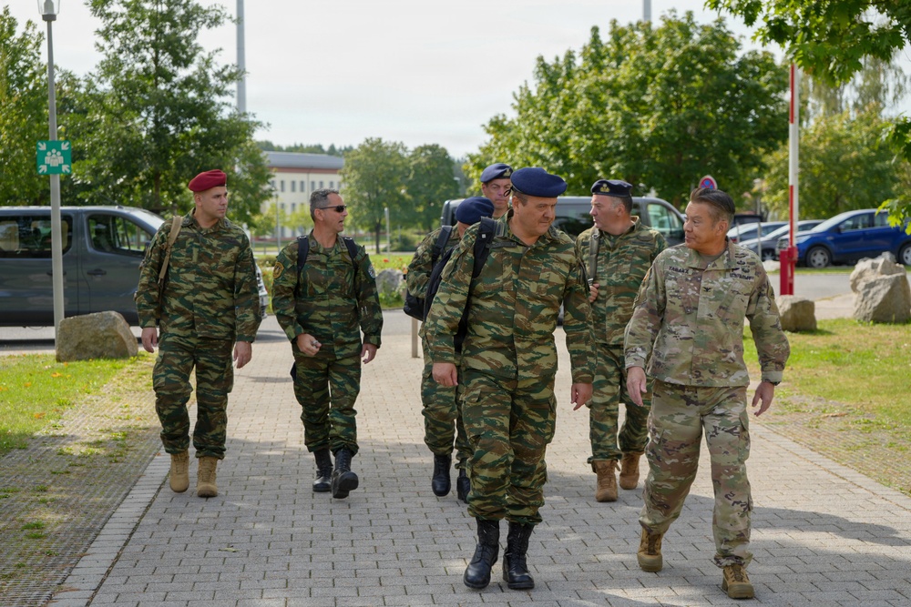 A group of military personnel from at least two different nations are walking together along a paved pathway, likely on a military installation or base. The majority wear a distinctive green lizard/tiger-stripe camouflage pattern with blue or red berets, consistent with Greek Armed Forces uniforms, while one individual on the right wears US Army MultiCam (OCP) pattern. The setting appears to be a European military base with trees, parked vehicles, and institutional buildings visible in the background.