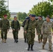 A group of military personnel from at least two different nations are walking together along a paved pathway, likely on a military installation or base. The majority wear a distinctive green lizard/tiger-stripe camouflage pattern with blue or red berets, consistent with Greek Armed Forces uniforms, while one individual on the right wears US Army MultiCam (OCP) pattern. The setting appears to be a European military base with trees, parked vehicles, and institutional buildings visible in the background.
