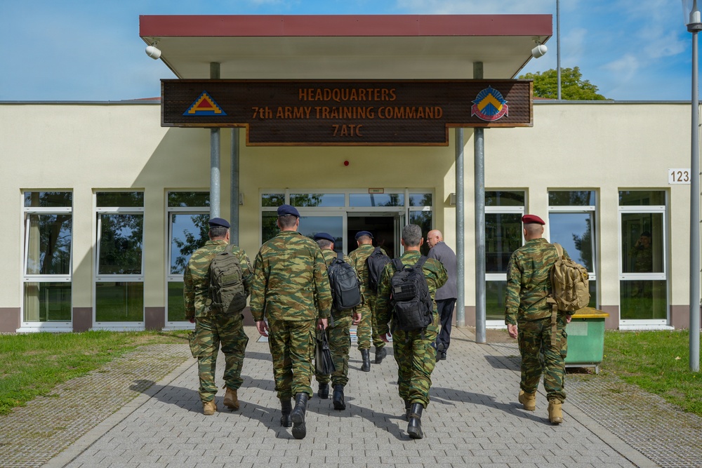 A group of military personnel in camouflage uniforms are walking toward the entrance of the Headquarters of the 7th Army Training Command (7ATC). The building displays a prominent wooden sign with the unit's name and insignia patches on either side. The scene appears to be at a military installation, likely in Europe based on the architectural style.