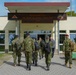 A group of military personnel in camouflage uniforms are walking toward the entrance of the Headquarters of the 7th Army Training Command (7ATC). The building displays a prominent wooden sign with the unit's name and insignia patches on either side. The scene appears to be at a military installation, likely in Europe based on the architectural style.