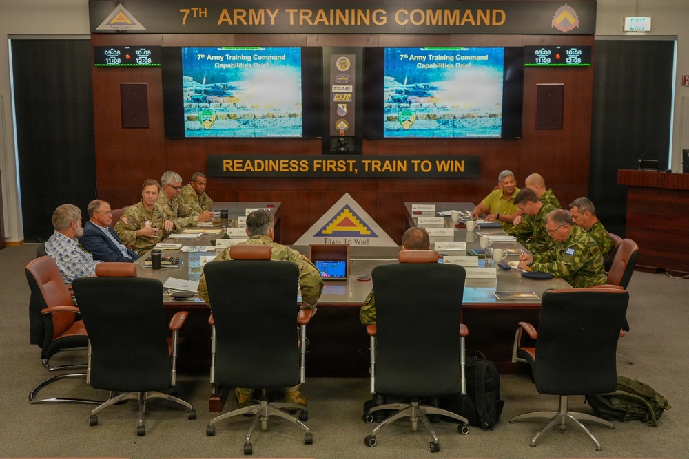 A formal briefing is taking place at the 7th Army Training Command headquarters, with military personnel and civilians seated around a large conference table. The room displays the command's motto 'Readiness First, Train To Win' and screens showing a '7th Army Training Command Capabilities Brief' presentation. Attendees include U.S. Army soldiers in ACU/OCP uniforms, individuals in foreign military camouflage uniforms, and civilian personnel.