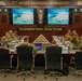 A formal briefing is taking place at the 7th Army Training Command headquarters, with military personnel and civilians seated around a large conference table. The room displays the command's motto 'Readiness First, Train To Win' and screens showing a '7th Army Training Command Capabilities Brief' presentation. Attendees include U.S. Army soldiers in ACU/OCP uniforms, individuals in foreign military camouflage uniforms, and civilian personnel.