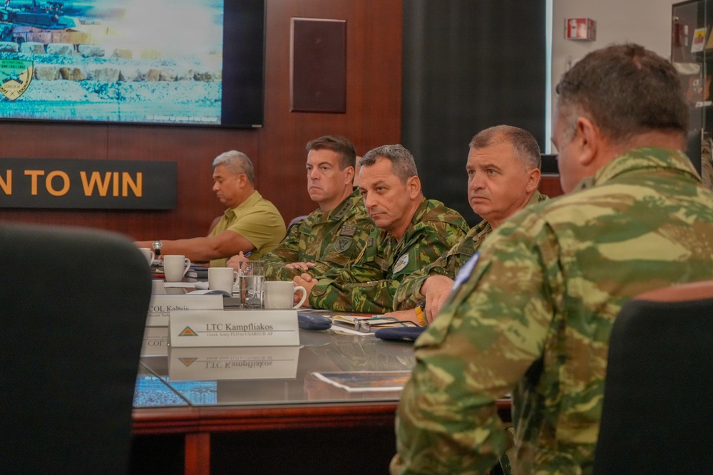 A military briefing or meeting is taking place in what appears to be a command center or conference room. Several military personnel in camouflage uniforms are seated at a conference table alongside at least one civilian. A display screen showing military imagery and a sign reading 'TO WIN' are visible in the background.