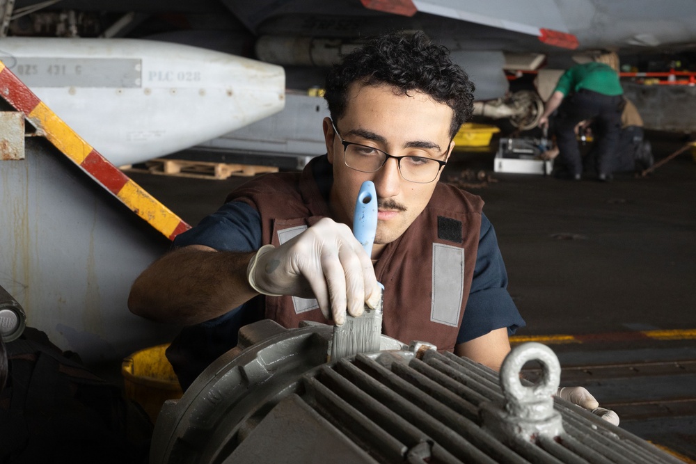 A young male technician wearing a brown vest and white latex gloves applies gray paint to a mechanical component using a brush in what appears to be an aircraft carrier hangar deck. A military aircraft is visible in the background, along with a second worker in a green shirt. The setting and equipment suggest a U.S. Navy maintenance operation.