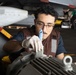 A young male technician wearing a brown vest and white latex gloves applies gray paint to a mechanical component using a brush in what appears to be an aircraft carrier hangar deck. A military aircraft is visible in the background, along with a second worker in a green shirt. The setting and equipment suggest a U.S. Navy maintenance operation.
