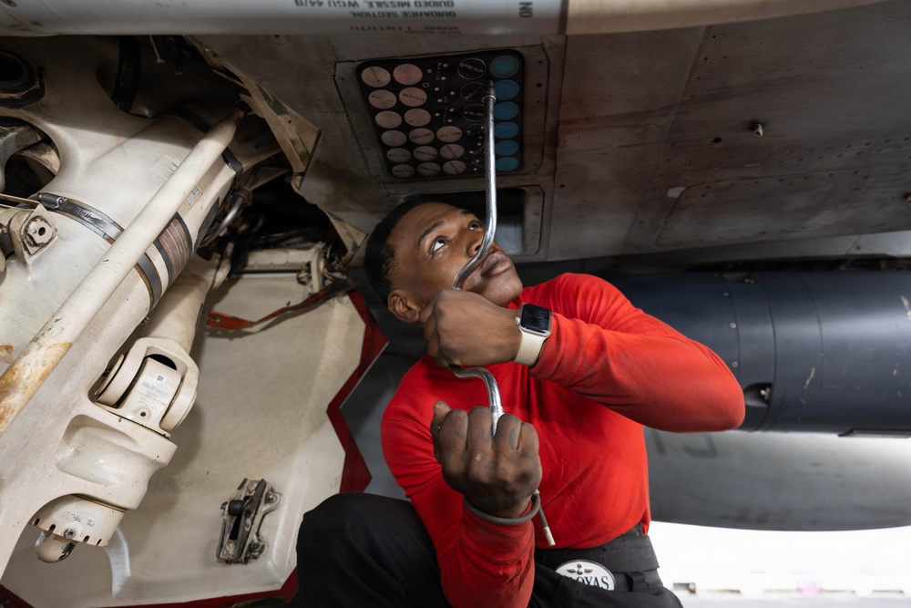 A technician wearing a red long-sleeve shirt is performing maintenance work underneath a military aircraft. He is using a ratchet/socket wrench tool and looking upward at the underside of the aircraft, focusing on a panel with multiple colored circular indicators or fasteners. The setting appears to be on a flight deck or maintenance area.