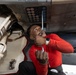 A technician wearing a red long-sleeve shirt is performing maintenance work underneath a military aircraft. He is using a ratchet/socket wrench tool and looking upward at the underside of the aircraft, focusing on a panel with multiple colored circular indicators or fasteners. The setting appears to be on a flight deck or maintenance area.