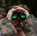 A person wearing digital camouflage military uniform is looking through black binoculars with distinctive green-coated lenses. The image is a close-up shot focused on the hands, binoculars, and lower face of the subject. At least one additional person in military attire is visible in the blurred background.