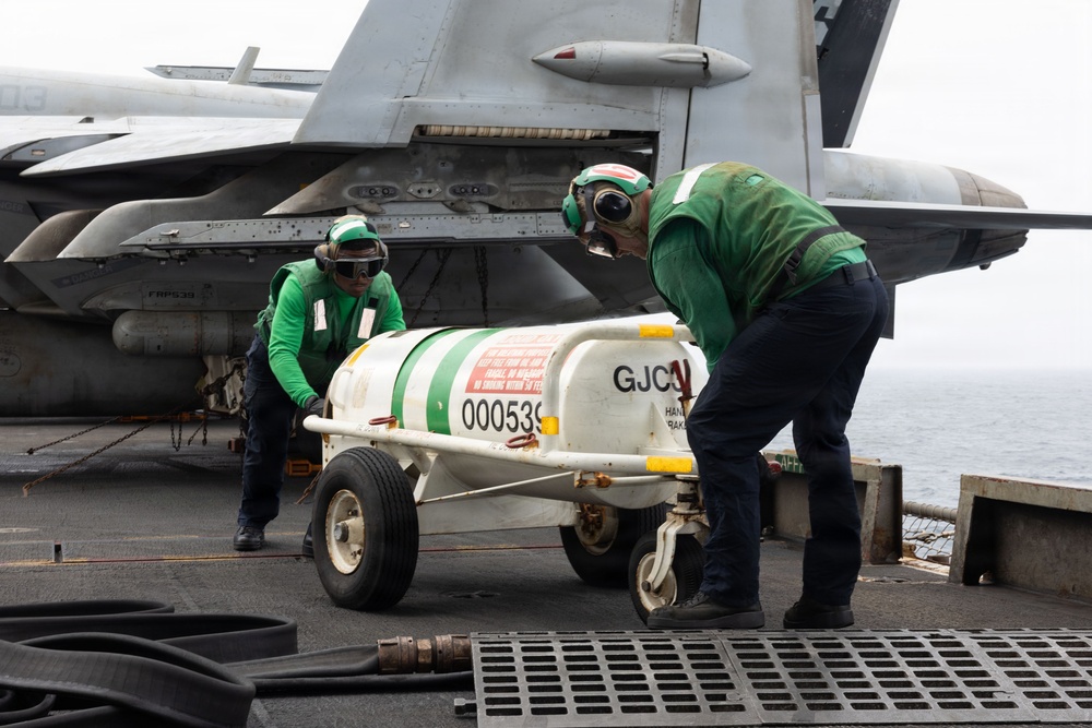 Two naval aviation personnel in green jerseys and helmets are handling a wheeled ordnance or equipment cart (marked 'GJC' and serial number '000539') on the flight deck of an aircraft carrier. A military jet aircraft, likely an F/A-18 Hornet or Super Hornet, is visible in the background. The scene depicts routine flight deck operations at sea.