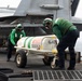 Two naval aviation personnel in green jerseys and helmets are handling a wheeled ordnance or equipment cart (marked 'GJC' and serial number '000539') on the flight deck of an aircraft carrier. A military jet aircraft, likely an F/A-18 Hornet or Super Hornet, is visible in the background. The scene depicts routine flight deck operations at sea.