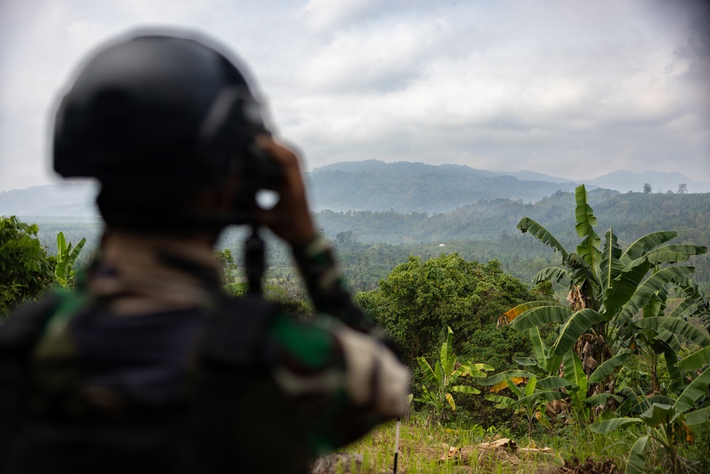 A soldier wearing a black helmet and camouflage uniform is using binoculars to observe a densely forested mountainous landscape. The scene appears to be a reconnaissance or surveillance operation conducted from an elevated vantage point. The surrounding vegetation, including banana trees and tropical foliage, suggests a Southeast Asian environment.