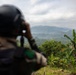 A soldier wearing a black helmet and camouflage uniform is using binoculars to observe a densely forested mountainous landscape. The scene appears to be a reconnaissance or surveillance operation conducted from an elevated vantage point. The surrounding vegetation, including banana trees and tropical foliage, suggests a Southeast Asian environment.