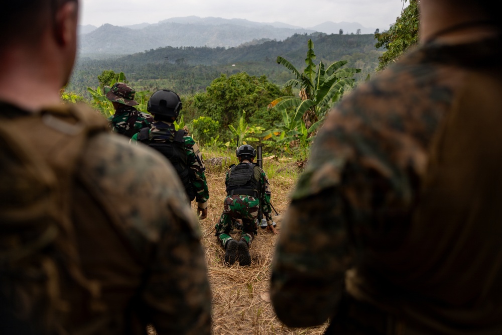 The image shows a group of military personnel in a tropical highland setting, with soldiers wearing two distinct camouflage patterns suggesting a joint or combined training exercise. One soldier is in a prone firing position with a weapon mounted on a bipod, while others observe from behind. The lush mountainous terrain with banana trees and layered ridgelines in the background suggests a Southeast Asian location.
