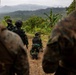 The image shows a group of military personnel in a tropical highland setting, with soldiers wearing two distinct camouflage patterns suggesting a joint or combined training exercise. One soldier is in a prone firing position with a weapon mounted on a bipod, while others observe from behind. The lush mountainous terrain with banana trees and layered ridgelines in the background suggests a Southeast Asian location.