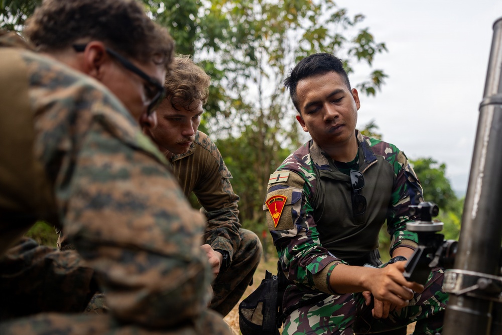 Three military personnel are gathered around what appears to be a mortar system in an outdoor, wooded setting. Two individuals wearing U.S. Marine Corps MARPAT camouflage uniforms are observing a third individual in a different camouflage pattern who appears to be demonstrating or operating the equipment. The scene suggests a joint military training exercise or equipment familiarization session.
