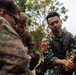 Three military personnel are gathered around what appears to be a mortar system in an outdoor, wooded setting. Two individuals wearing U.S. Marine Corps MARPAT camouflage uniforms are observing a third individual in a different camouflage pattern who appears to be demonstrating or operating the equipment. The scene suggests a joint military training exercise or equipment familiarization session.