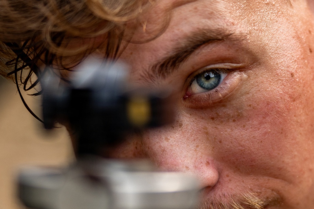 An extreme close-up photograph of a young man with blue eyes, freckles, and curly reddish-blonde hair. He appears to be looking through or alongside a scope or optical sighting device, which is blurred in the foreground. The shallow depth of field creates a sharp focus on his right eye and facial features.