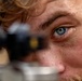 An extreme close-up photograph of a young man with blue eyes, freckles, and curly reddish-blonde hair. He appears to be looking through or alongside a scope or optical sighting device, which is blurred in the foreground. The shallow depth of field creates a sharp focus on his right eye and facial features.