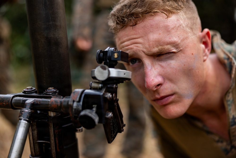 A young male soldier in camouflage uniform is looking through the sight of what appears to be a mortar or artillery aiming device. He has short blonde hair, visible face paint/camouflage markings, and is squinting with one eye closed to focus through the optical sight. The setting appears to be an outdoor wooded or jungle training environment.