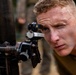 A young male soldier in camouflage uniform is looking through the sight of what appears to be a mortar or artillery aiming device. He has short blonde hair, visible face paint/camouflage markings, and is squinting with one eye closed to focus through the optical sight. The setting appears to be an outdoor wooded or jungle training environment.