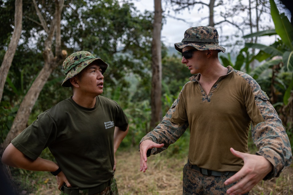 Two military personnel in camouflage uniforms are engaged in conversation in an outdoor jungle or forested environment. One individual appears to be of Asian descent wearing an olive green t-shirt with a camouflage bucket hat, while the other wears a full MARPAT (Marine Pattern) uniform with a matching boonie hat and sunglasses. The taller individual appears to be gesturing with his hands, suggesting an active briefing or discussion.