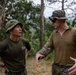 Two military personnel in camouflage uniforms are engaged in conversation in an outdoor jungle or forested environment. One individual appears to be of Asian descent wearing an olive green t-shirt with a camouflage bucket hat, while the other wears a full MARPAT (Marine Pattern) uniform with a matching boonie hat and sunglasses. The taller individual appears to be gesturing with his hands, suggesting an active briefing or discussion.