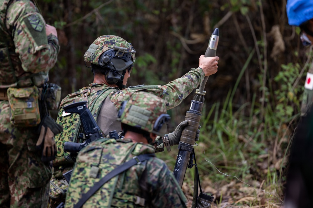Military personnel in camouflage uniforms are operating what appears to be a mortar system in a wooded outdoor environment. One soldier is holding a mortar round above the tube, appearing to demonstrate or prepare for a loading procedure. The uniforms and equipment are consistent with Japan Ground Self-Defense Force (JGSDF) pattern and gear.