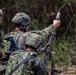 Military personnel in camouflage uniforms are operating what appears to be a mortar system in a wooded outdoor environment. One soldier is holding a mortar round above the tube, appearing to demonstrate or prepare for a loading procedure. The uniforms and equipment are consistent with Japan Ground Self-Defense Force (JGSDF) pattern and gear.