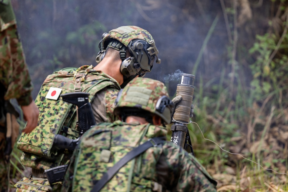 Two soldiers from the Japan Ground Self-Defense Force (JGSDF) are visible in a field/woodland environment during what appears to be a military training exercise. One soldier is holding a smoking device, likely a smoke grenade or similar pyrotechnic, while smoke is visible in the background. Both soldiers are wearing standard JGSDF camouflage uniforms with tactical gear.