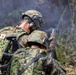 Two soldiers from the Japan Ground Self-Defense Force (JGSDF) are visible in a field/woodland environment during what appears to be a military training exercise. One soldier is holding a smoking device, likely a smoke grenade or similar pyrotechnic, while smoke is visible in the background. Both soldiers are wearing standard JGSDF camouflage uniforms with tactical gear.